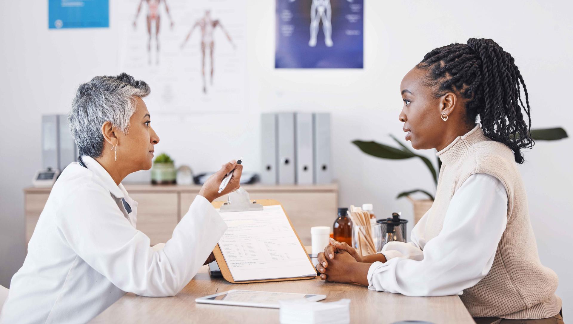 Two women in a sugerical office setting have a conversation, seated at a table